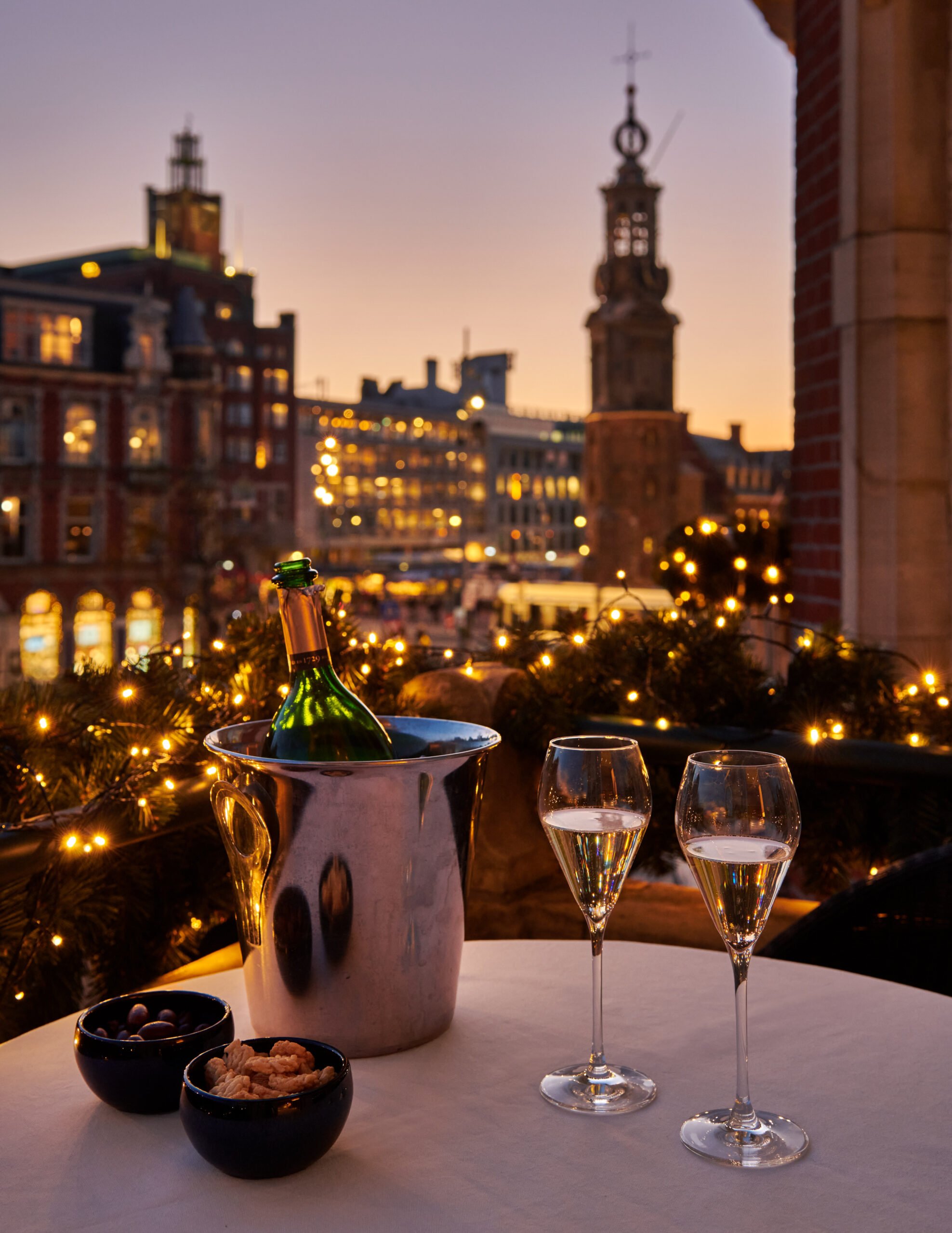 Nighttime scene with festive Christmas lighting, two glasses of champagne and a bottle chilling in ice, overlooking the illuminated Munt Tower.